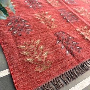 May include: Red woven rug with a geometric pattern of brown and beige leaves. The rug has a fringe border.