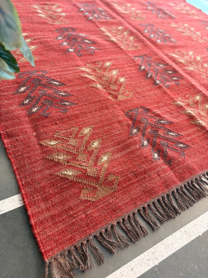 May include: Red woven rug with a geometric pattern of brown and beige leaves. The rug has a fringe border.