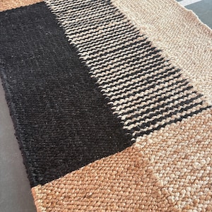 Può includere: Un tappeto rettangolare in juta con un motivo geometrico bianco e nero. Il tappeto ha un tono naturale e terroso ed è tessuto con un aspetto testurizzato e artigianale.