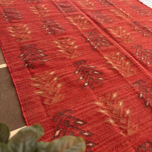 May include: Red, brown, and beige patterned rug with a woven texture. The rug has a geometric design with a repeating pattern of stylized leaves.