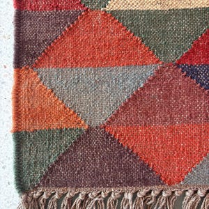May include: A geometric patterned rug with a fringe border. The rug is made of woven fabric in shades of red, orange, brown, green, and grey.