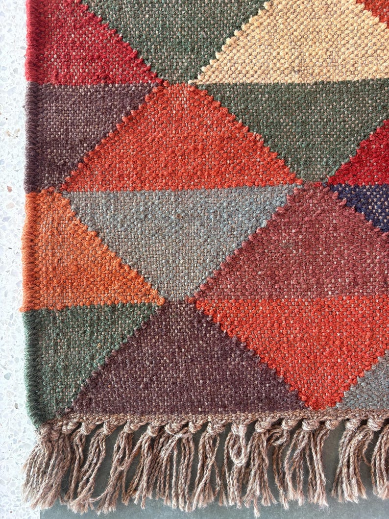 May include: A geometric patterned rug with a fringe border. The rug is made of woven fabric in shades of red, orange, brown, green, and grey.