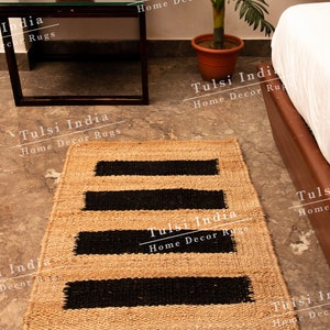 May include: A natural fibre rug with black stripes woven into the beige material. The rug is on a light grey floor with a small black metal table and a potted plant in the background. The text "Tulsi India Home Decor Rugs" is repeated throughout the image.