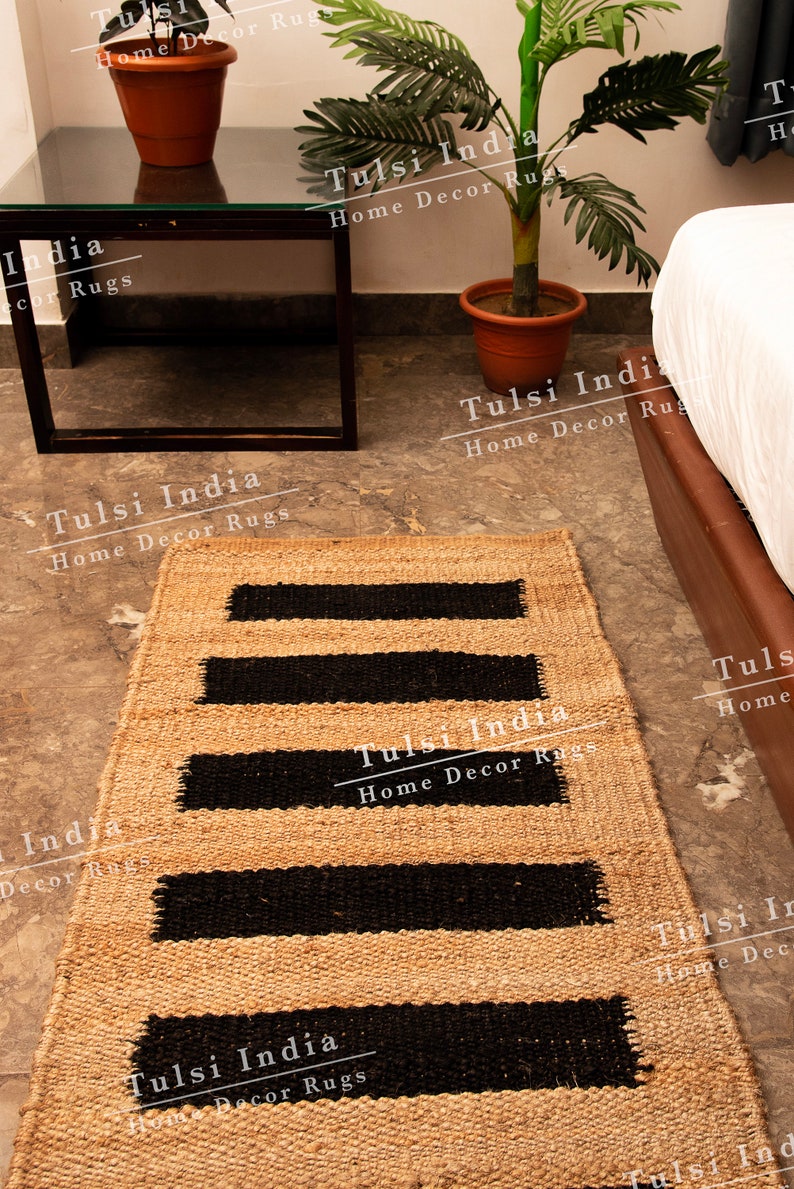 May include: A natural fibre rug with black stripes woven into the beige material. The rug is on a light grey floor with a small black metal table and a potted plant in the background. The text "Tulsi India Home Decor Rugs" is repeated throughout the image.