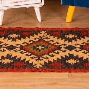 Puede incluir: Una alfombra roja, negra y amarilla con flecos en los extremos. La alfombra tiene un dise&ntilde;o geom&eacute;trico con formas de diamantes y un estilo tribal.