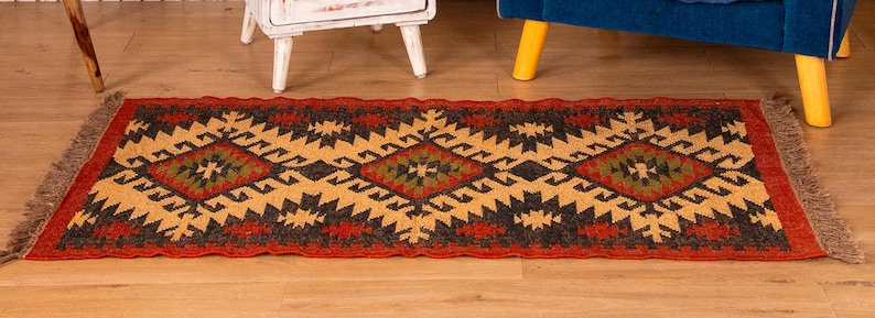 Puede incluir: Una alfombra roja, negra y amarilla con flecos en los extremos. La alfombra tiene un dise&ntilde;o geom&eacute;trico con formas de diamantes y un estilo tribal.