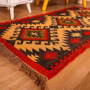 Puede incluir: Una alfombra colorida con estampado geom&eacute;trico y flecos en los bordes. La alfombra es en tonos rojos, negros, amarillos y verdes.