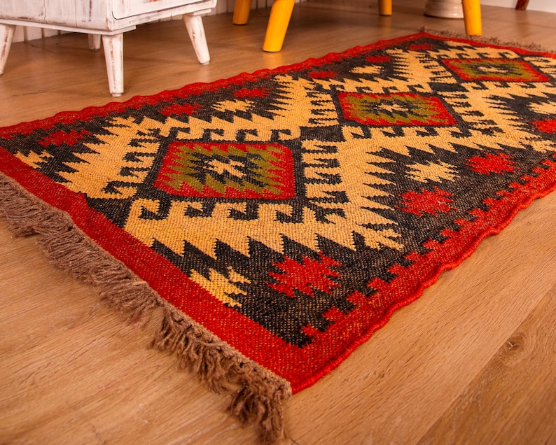 Puede incluir: Una alfombra colorida con estampado geom&eacute;trico y flecos en los bordes. La alfombra es en tonos rojos, negros, amarillos y verdes.