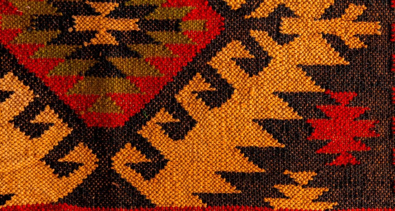 Puede incluir: Primer plano de una alfombra tejida con un patr&oacute;n geom&eacute;trico en tonos rojos, naranjas, negros y verdes.