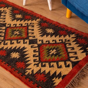 Puede incluir: Una alfombra de estilo tradicional con un patr&oacute;n geom&eacute;trico en tonos rojos, negros, verdes y beige. La alfombra tiene un borde de flecos.