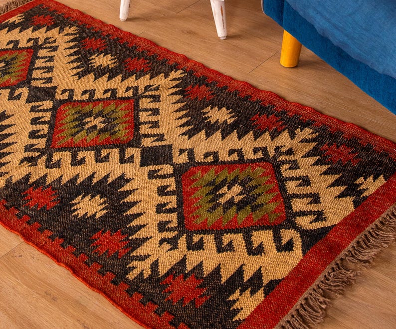 Puede incluir: Una alfombra de estilo tradicional con un patr&oacute;n geom&eacute;trico en tonos rojos, negros, verdes y beige. La alfombra tiene un borde de flecos.