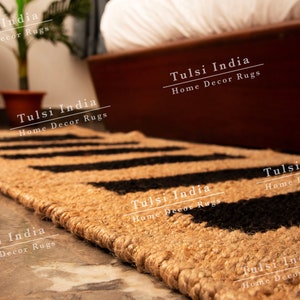 May include: A close-up of a natural fibre rug with a black and tan geometric pattern. The rug is made of jute and has a fringed edge. The rug is labelled "Tulsi India Home Decor Rugs".