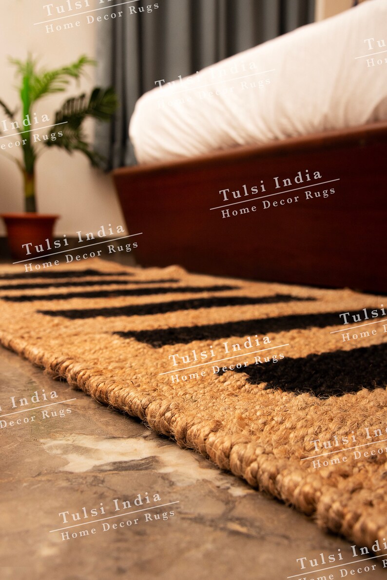 May include: A close-up of a natural fibre rug with a black and tan geometric pattern. The rug is made of jute and has a fringed edge. The rug is labelled "Tulsi India Home Decor Rugs".