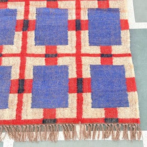 May include: A woven rug with a geometric pattern of red, blue, and white squares. The rug has a fringe border.
