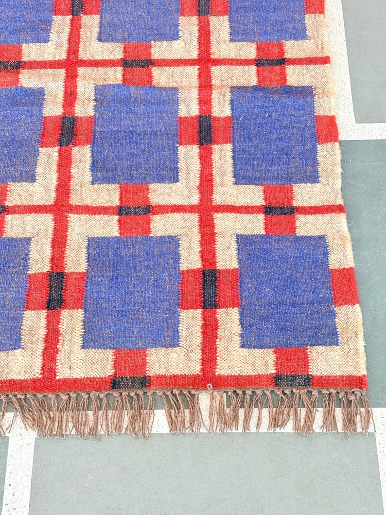May include: A woven rug with a geometric pattern of red, blue, and white squares. The rug has a fringe border.