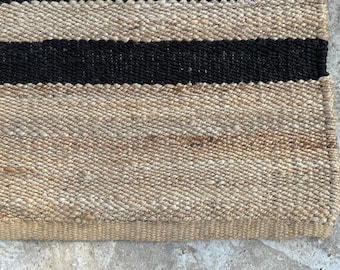 May include: Close-up of a woven jute rug with a natural beige background and two thick black stripes. The rug has a textured surface and a woven border.
