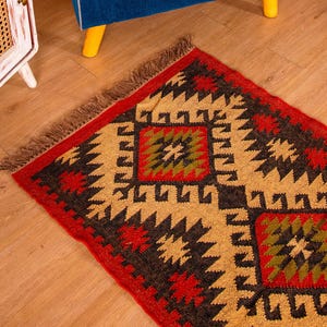 Puede incluir: Una alfombra roja, negra y beige con flecos. La alfombra tiene un dise&ntilde;o geom&eacute;trico con una forma de diamante en el centro.
