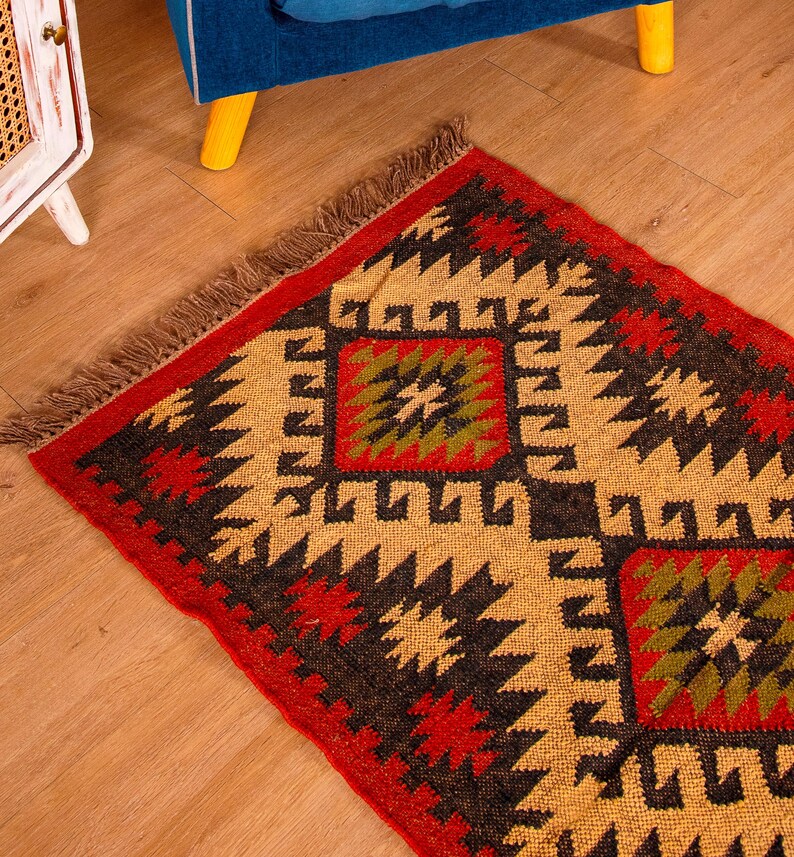 Puede incluir: Una alfombra roja, negra y beige con flecos. La alfombra tiene un dise&ntilde;o geom&eacute;trico con una forma de diamante en el centro.