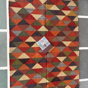 May include: A colourful geometric rug with a pattern of triangles in shades of red, orange, brown, green, and beige. The rug has a fringe border.