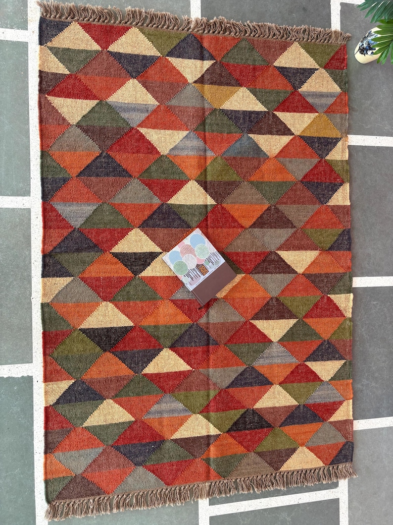 May include: A colourful geometric rug with a pattern of triangles in shades of red, orange, brown, green, and beige. The rug has a fringe border.