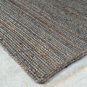 Può includere: Primo piano di un tappeto rettangolare in juta intrecciata. Il tappeto presenta un mix di tonalità grigie, marroni e beige, creando un aspetto strutturato. Il motivo intrecciato è visibile e i bordi sono rifiniti con cura. Questo tappeto è adatto per l'arredamento della casa.