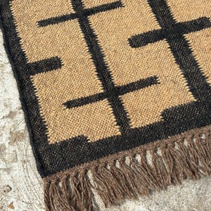 May include: Close-up of a woven rug with a geometric pattern. The rug features a tan background with a repeating black design. The edge of the rug has a black border and brown fringe.