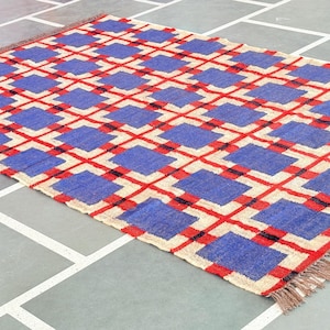 May include: A blue and red geometric patterned rug with fringe on the edges. The rug is made of woven fibers and has a cream background.