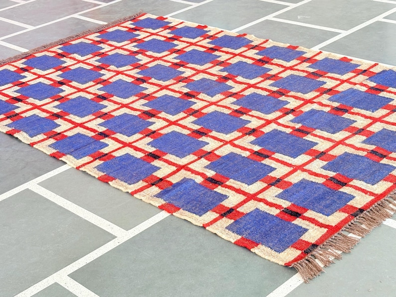 May include: A blue and red geometric patterned rug with fringe on the edges. The rug is made of woven fibers and has a cream background.