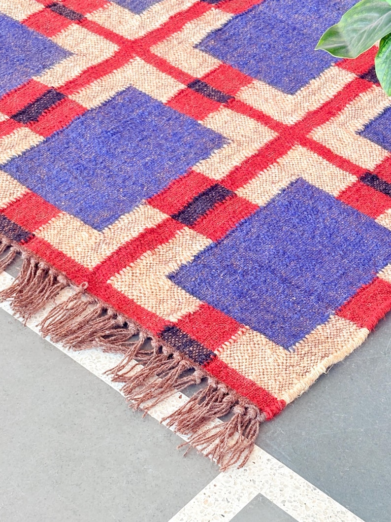 May include: A woven rug with a geometric pattern in shades of blue, red, and beige. The rug has a fringe border.