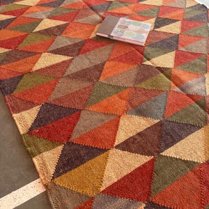 May include: A multi-coloured geometric patterned rug with a woven texture. The rug features a variety of shades of brown, red, orange, and green. The rug is made of natural fibres and has a fringed edge.