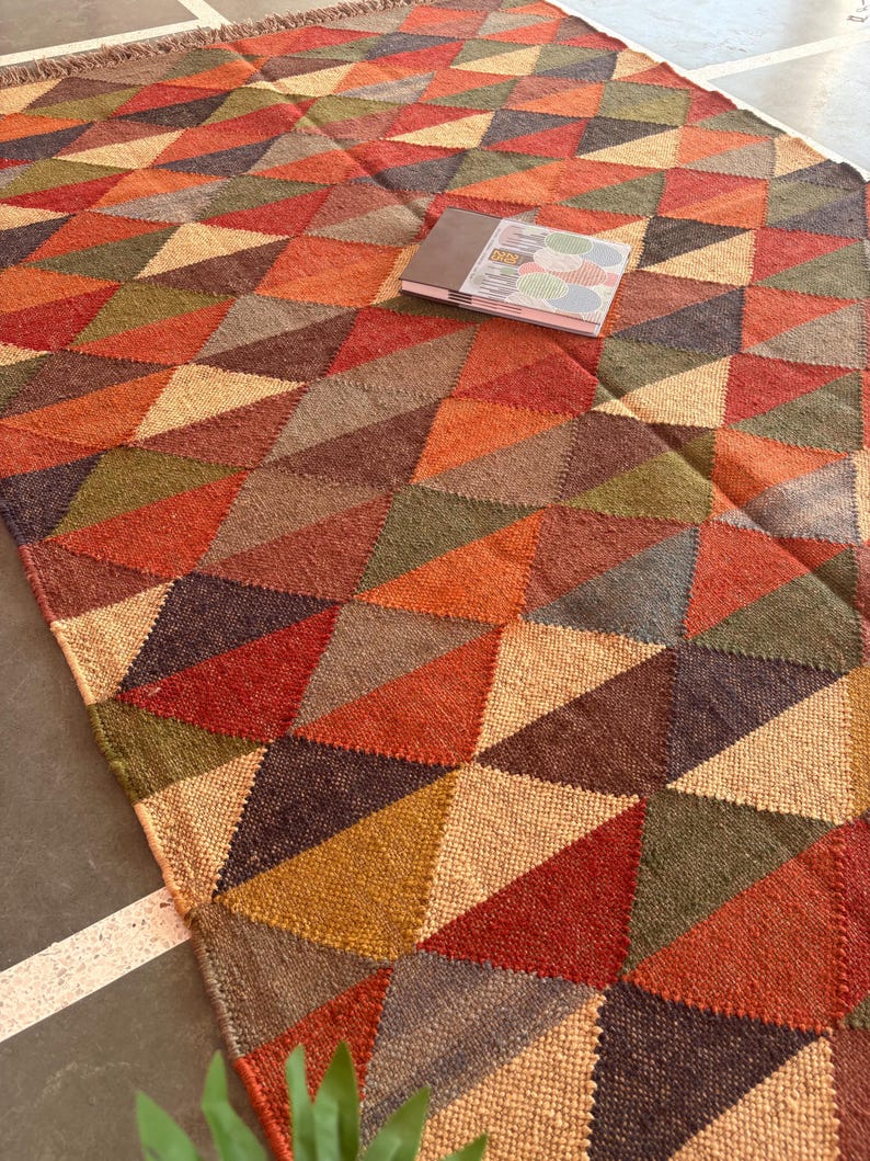 May include: A multi-coloured geometric patterned rug with a woven texture. The rug features a variety of shades of brown, red, orange, and green. The rug is made of natural fibres and has a fringed edge.