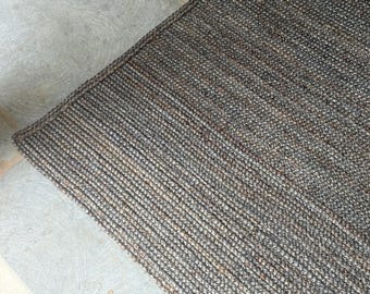 May include: A rectangular rug with a woven texture in shades of gray and brown. The rug's edges are neatly finished with a darker border. The rug is made of natural fibers and is suitable for indoor use.