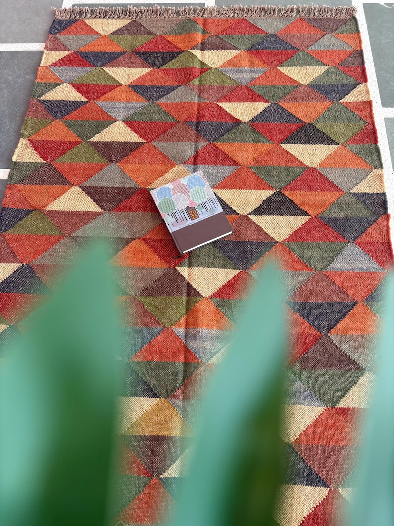 May include: A colourful rug with a geometric pattern of triangles in shades of red, orange, green, brown, and beige. The rug has a fringed border and is made of a woven material.
