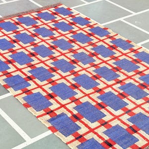 May include: A woven rug with a geometric pattern of red, blue, and white squares. The rug has a fringe border.