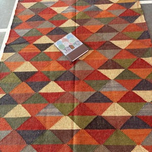 May include: A multicoloured geometric patterned rug with a fringe border. The rug is made of woven jute and features a variety of shades of brown, red, green, and beige.