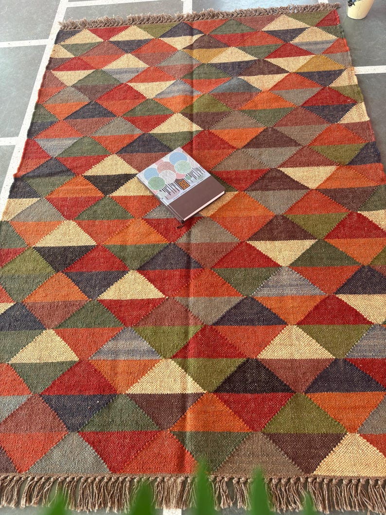 May include: A multicoloured geometric patterned rug with a fringe border. The rug is made of woven jute and features a variety of shades of brown, red, green, and beige.