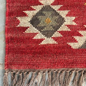 Könnte beinhalten: Nahaufnahme eines roten gewebten Teppichs mit einem geometrischen Rautenmuster in Weiß, Grau und Gelb. Der Teppich hat einen gefransten Rand in einer neutralen braunen Farbe. Der Teppich besteht aus einem strukturierten Material.