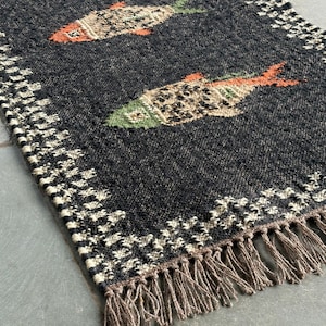 May include: A rectangular woven rug in dark charcoal gray featuring two fish motifs with orange and green accents. The rug has a textured border with a repeating cream and black pattern and is finished with a fringe of brown tassels.