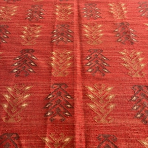 May include: Red rug with a geometric pattern of brown and gold stylized trees. The rug has a woven texture.