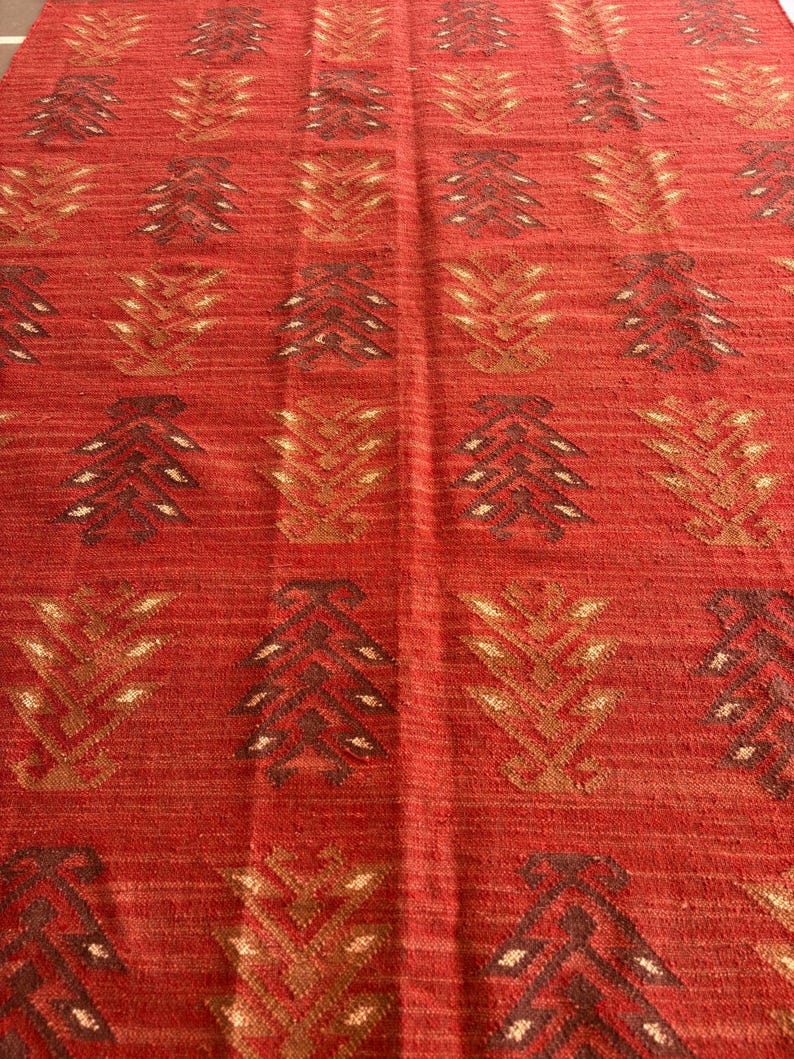 May include: Red rug with a geometric pattern of brown and gold stylized trees. The rug has a woven texture.