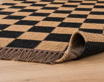 May include: A rectangular area rug featuring a repeating black and tan checkered pattern. The rug is constructed from a coarse, natural fiber material and is finished with a row of short, thin, brown fringe along the bottom edge.