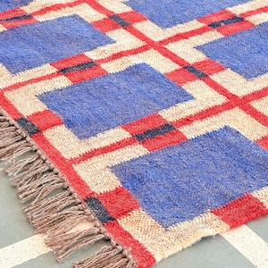 May include: A woven rug with a geometric pattern of red, blue, and beige squares. The rug has fringe on all sides.