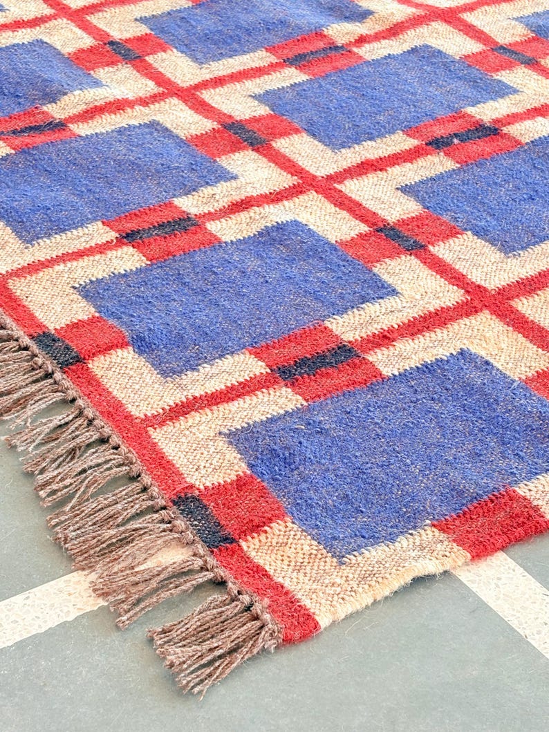 May include: A woven rug with a geometric pattern of red, blue, and beige squares. The rug has fringe on all sides.