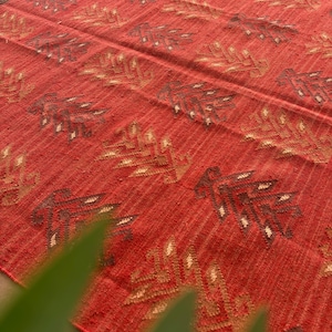 May include: Red, brown, and beige woven rug with a geometric pattern. The rug has a textured surface and a fringe border.