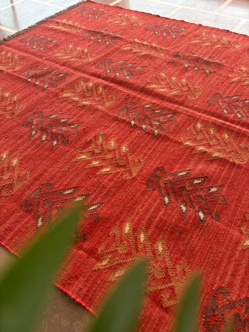 May include: Red, brown, and beige woven rug with a geometric pattern. The rug has a textured surface and a fringe border.