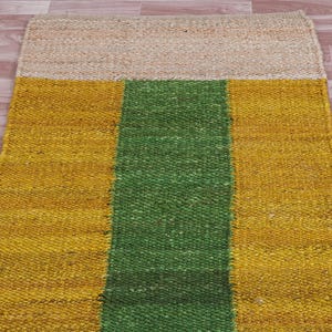 Pode incluir: Um tapete retangular de juta com uma faixa verde no centro e faixas amarelas de cada lado. O tapete tem uma textura natural e tecida.