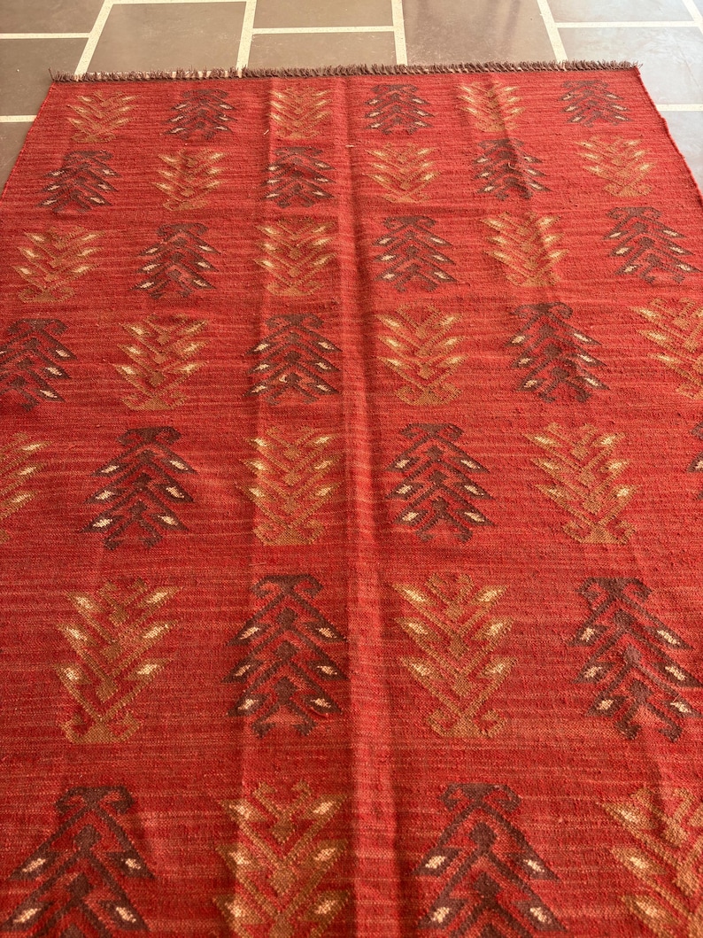 May include: Red area rug with a geometric pattern of brown and gold stylized leaves. The rug has a fringe border.