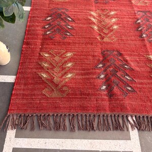 May include: Red and brown patterned rug with fringe. The rug has a geometric design with a repeating pattern of stylized trees.