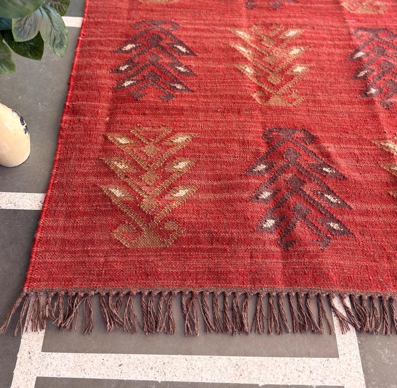 May include: Red and brown patterned rug with fringe. The rug has a geometric design with a repeating pattern of stylized trees.