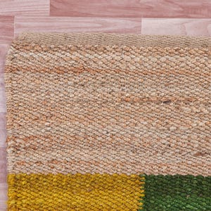 Pode incluir: Um tapete tecido com uma se&ccedil;&atilde;o superior bege natural e uma se&ccedil;&atilde;o inferior com tr&ecirc;s listras horizontais em amarelo, verde e marrom.